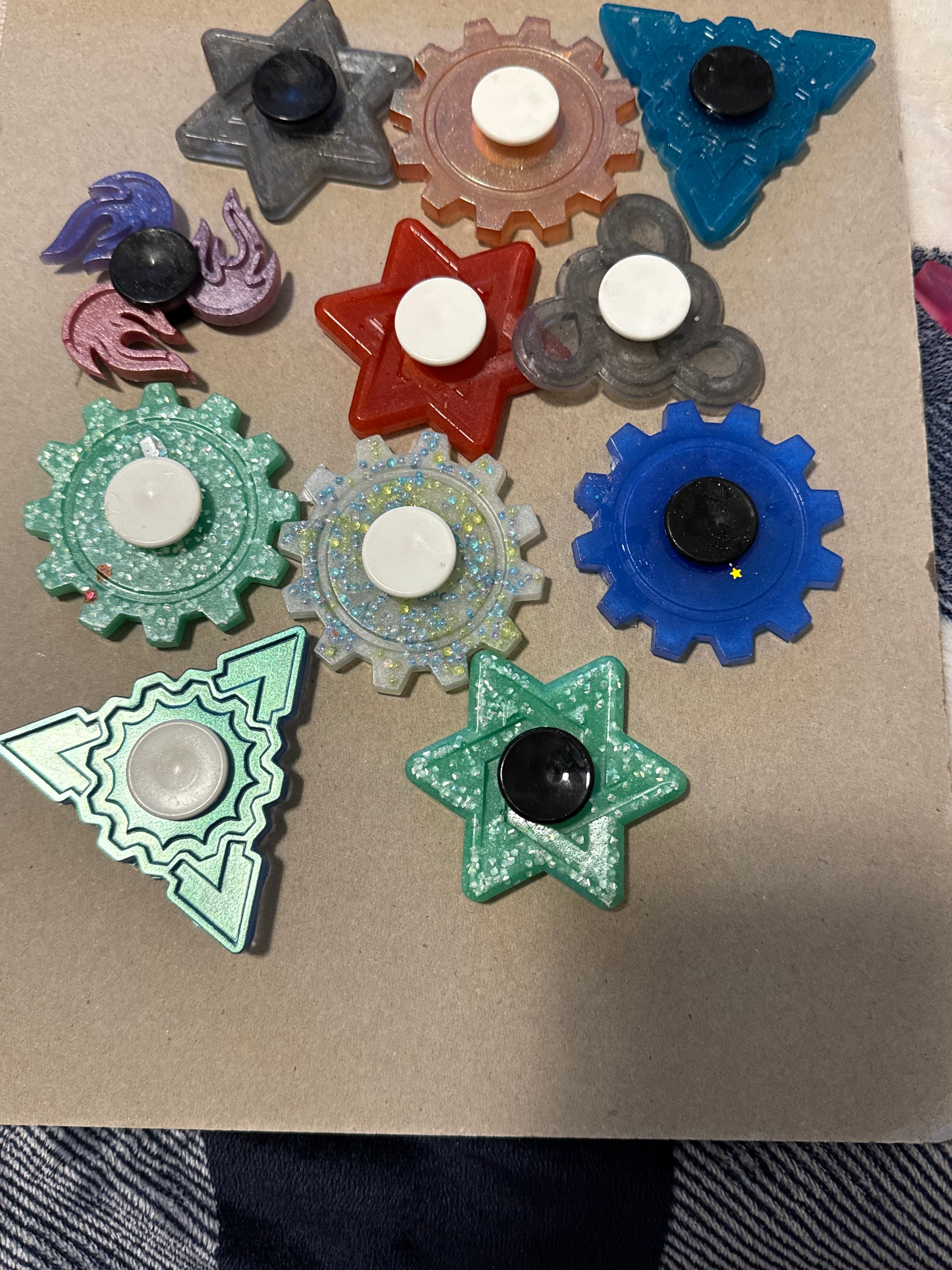 A set of colorful plastic fidget spinner toys arranged on a tan cardboard surface. The set includes various shapes like stars, triangles, and gears in blue, green, red, purple, and clear plastic, with black and white center buttons.